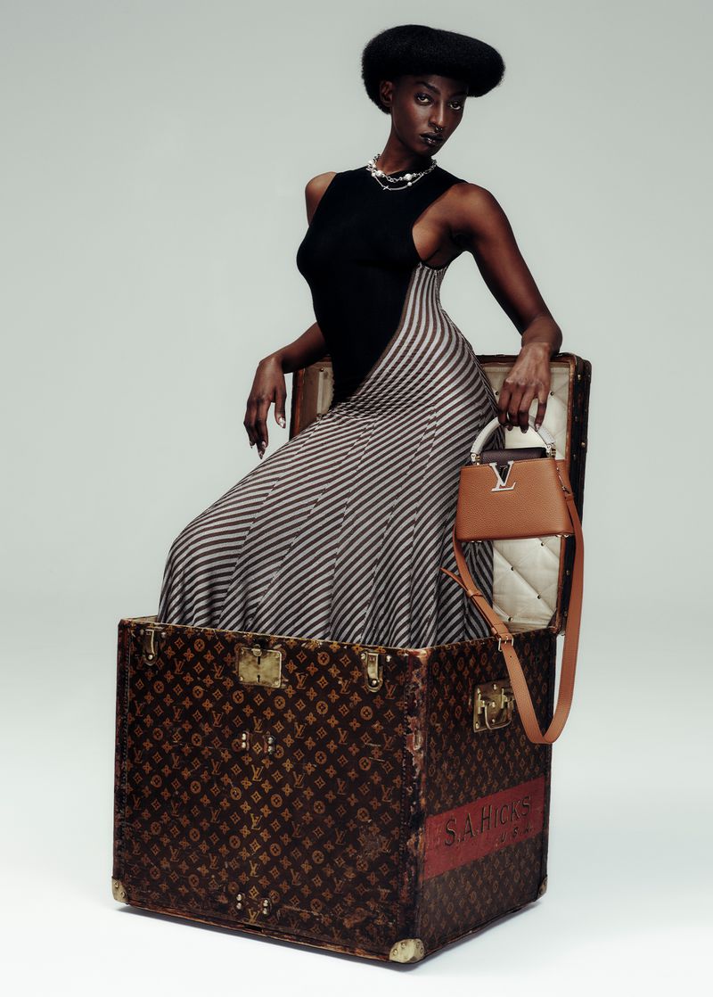 A Black woman poses in a striped dress and black top while sitting in a Louis Vuitton trunk with a handbag on a white background.
