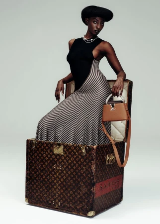 A Black woman poses in a striped dress and black top while sitting in a Louis Vuitton trunk with a handbag on a white background.