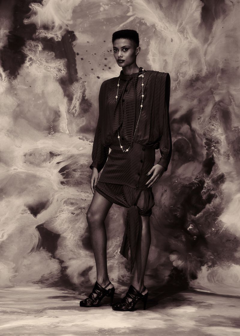 Monochrome fashion editorial portrait of a woman standing against a swirling textured backdrop, wearing a dark patterned dress and heels.