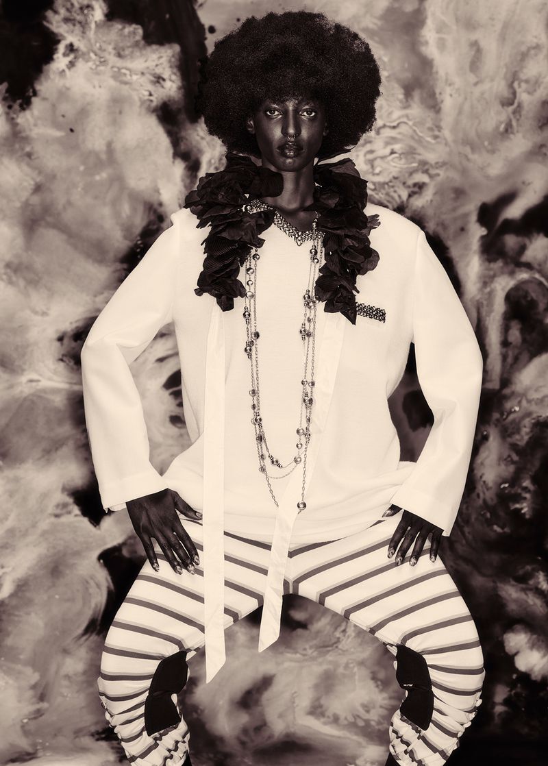 Model with large afro hairstyle wearing white top and striped trousers against a marbled abstract background.