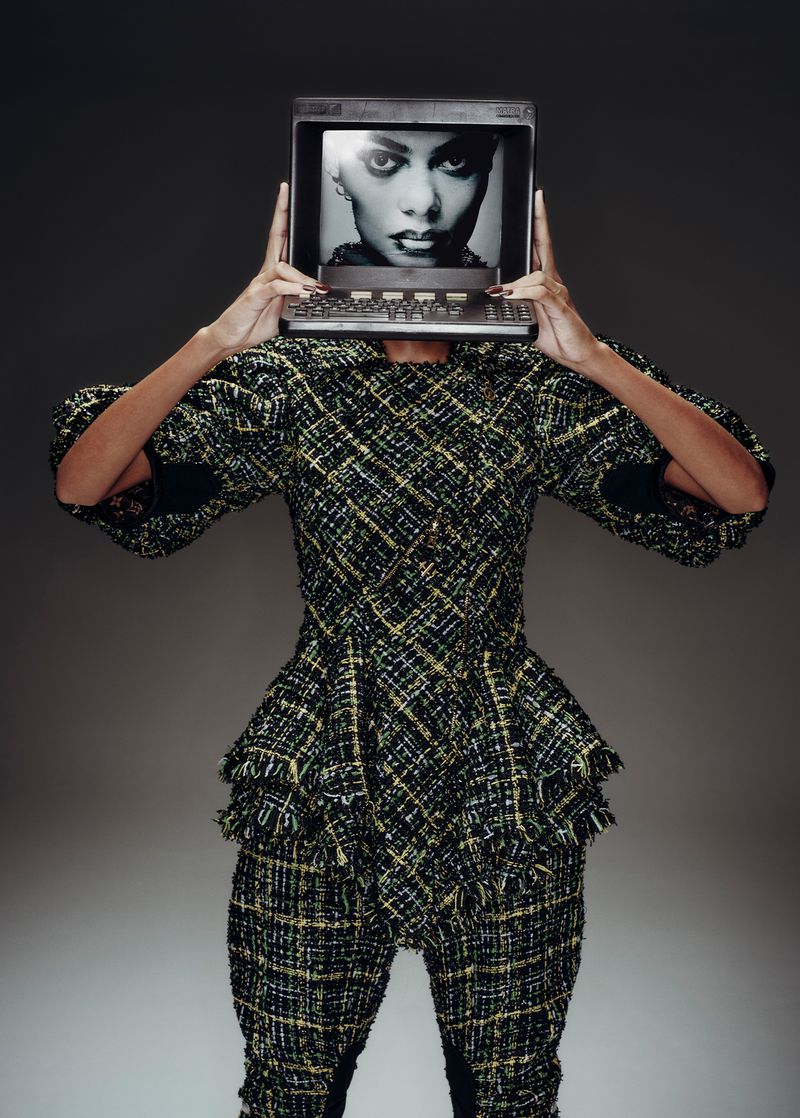Model in tweed suit holds computer monitor with fashion portrait in front of face against grey background in studio shot.
