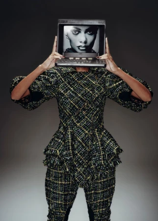 Model in tweed suit holds computer monitor with fashion portrait in front of face against grey background in studio shot.