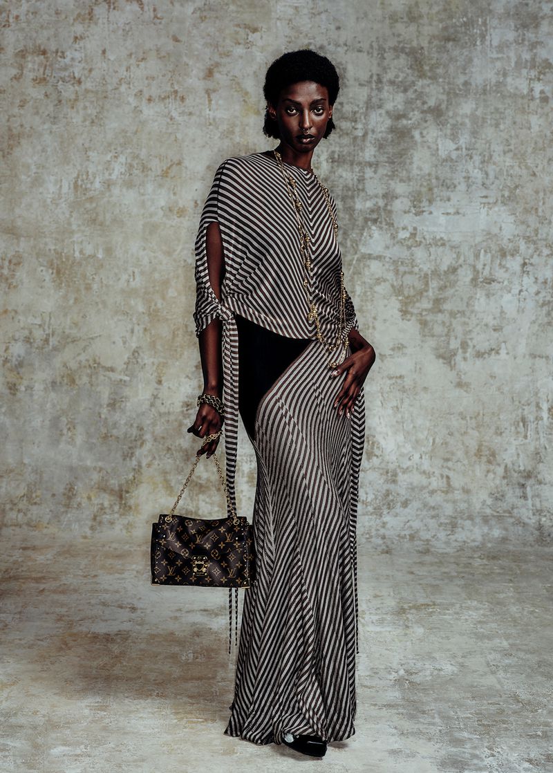 A slender, dark-skinned woman in a striped dress holds a Louis Vuitton purse in a fashion studio setting, illuminated by soft lighting.