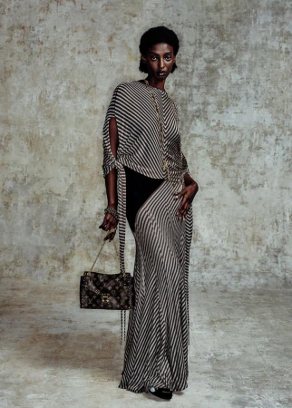 A slender, dark-skinned woman in a striped dress holds a Louis Vuitton purse in a fashion studio setting, illuminated by soft lighting.