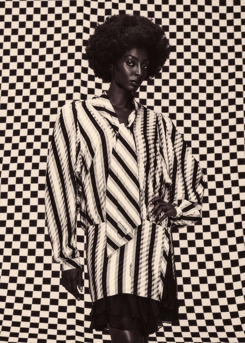 Black woman with an afro wearing a striped long-sleeve dress against a checkered backdrop in a monochrome fashion editorial portrait.