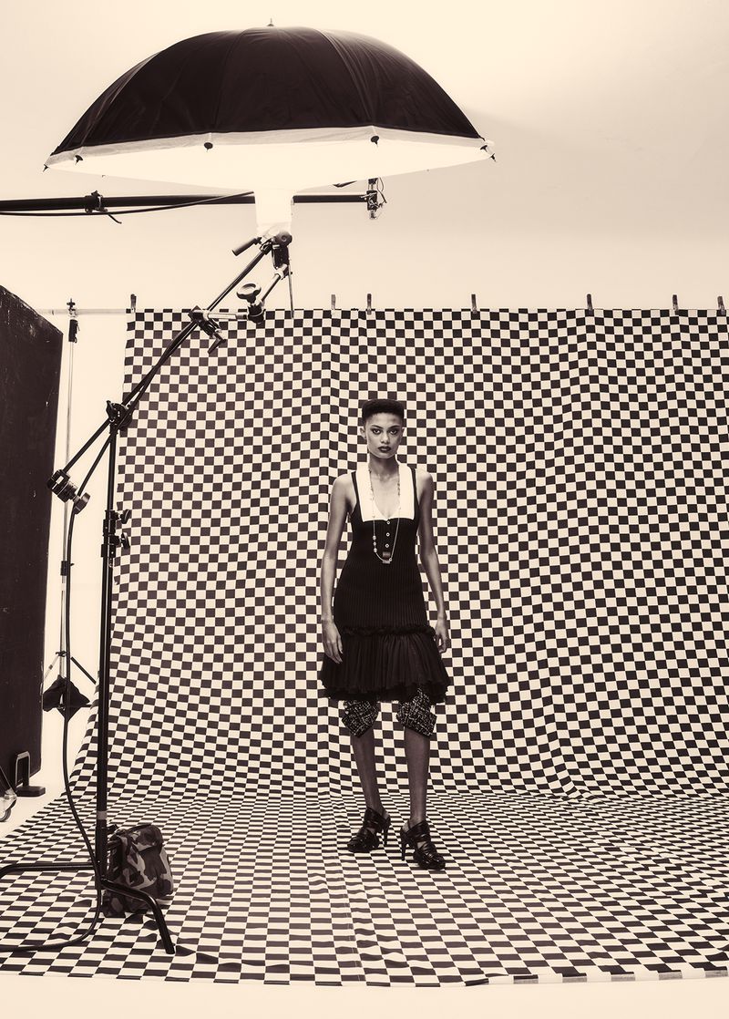 Fashion photo of a woman in black dress and high heels standing in a studio with a checkered backdrop and lighting equipment.