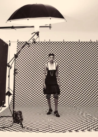 Fashion photo of a woman in black dress and high heels standing in a studio with a checkered backdrop and lighting equipment.