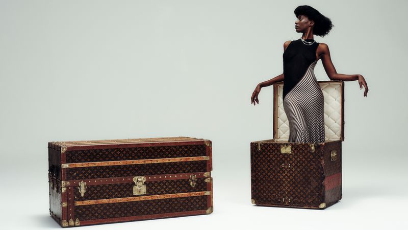 An elegant Black woman stands in a vintage Louis Vuitton trunk next to another matching trunk against a solid gray background.