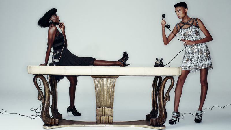 Two women in dresses pose with vintage telephones on an ornate table against a light backdrop, creating a stylish fashion tableau.
