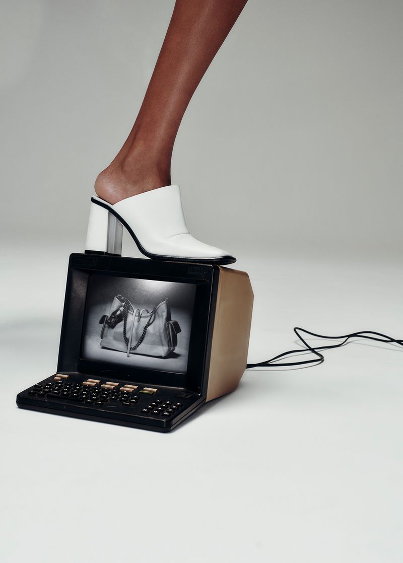 A person's leg and foot, wearing a white high-heeled shoe, is resting on a vintage computer monitor against a white background.
