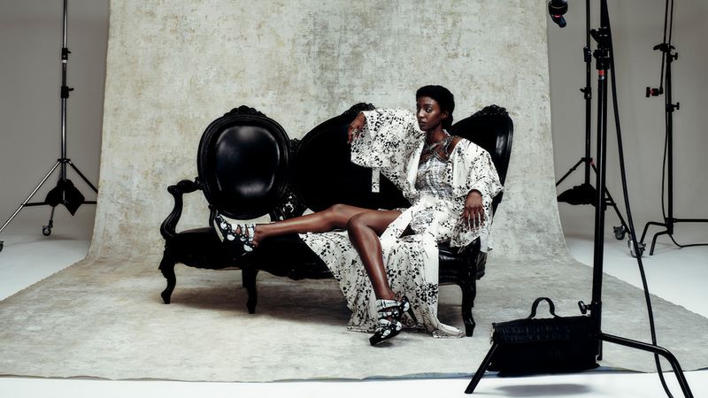 Elegant Black woman in patterned robe and shoes poses on a black vintage couch in a photo studio with lighting equipment.