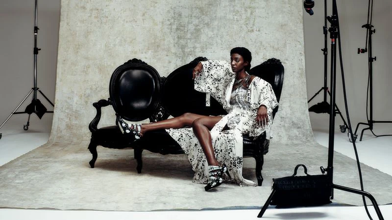Elegant Black woman in patterned robe and shoes poses on a black vintage couch in a photo studio with lighting equipment.