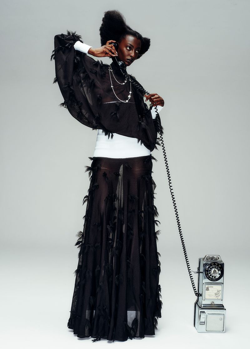A tall, slender, adult female fashion model poses with an antique telephone in a light studio setting, wearing a sheer black feather dress.