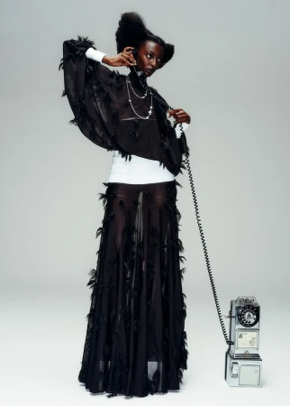 A tall, slender, adult female fashion model poses with an antique telephone in a light studio setting, wearing a sheer black feather dress.