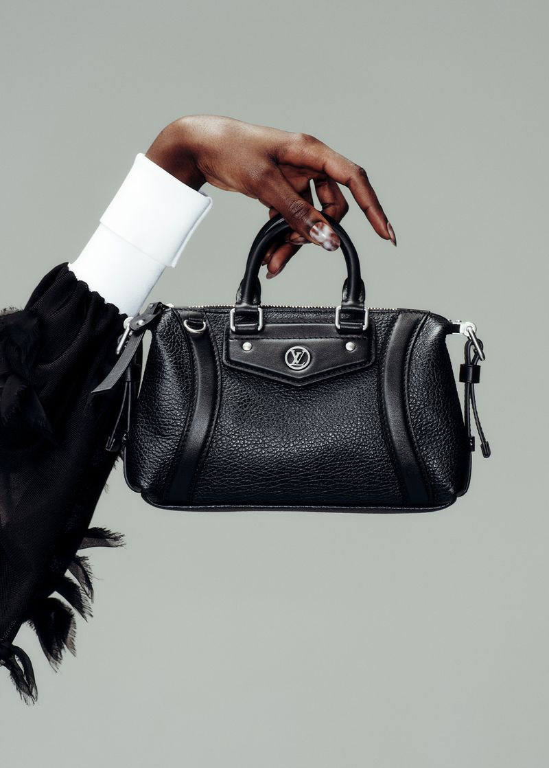 Close-up of a Black leather handbag with a logo, held by a dark-skinned hand with painted nails; grey backdrop, black clothing and white cuff visible.