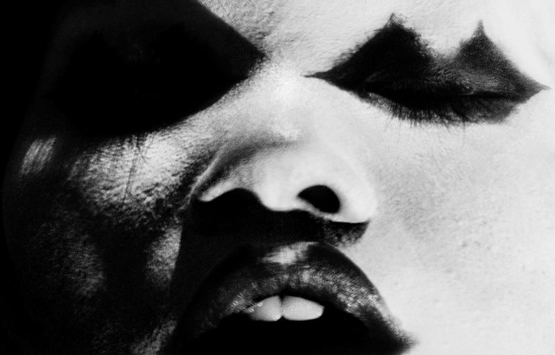 Extreme close-up portrait of a person with high-contrast black and white makeup, dark heavy eye shadow, and parted lips.