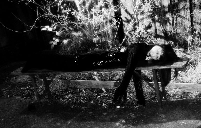 Monochrome image of a person lying on a wooden bench outdoors with branches and leaves in the background in a dark and artistic scene.