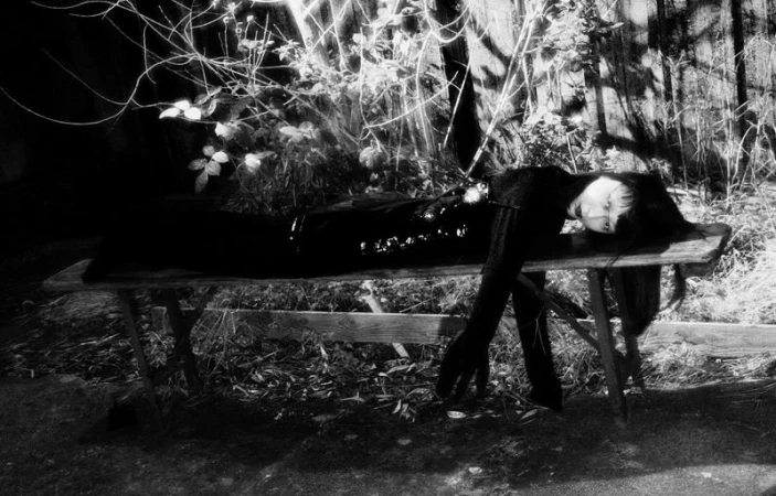 Monochrome image of a person lying on a wooden bench outdoors with branches and leaves in the background in a dark and artistic scene.