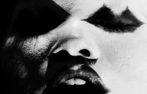 Extreme close-up portrait of a person with high-contrast black and white makeup, dark heavy eye shadow, and parted lips.