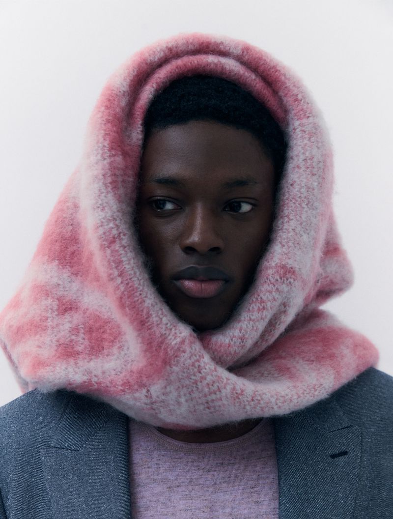 Portrait of a young Black man in a grey jacket with a pink knit shirt and pink and white patterned knit headscarf against white backdrop.