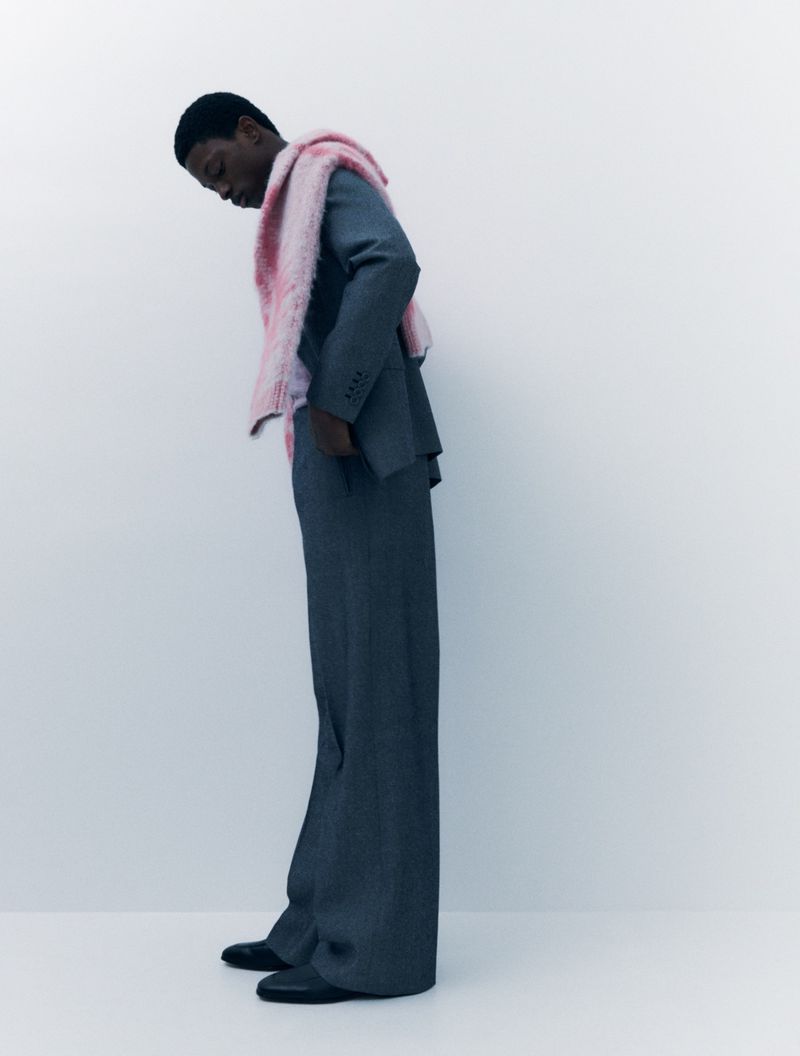 A slender Black male model poses indoors wearing a tailored grey suit, wide-legged trousers, black shoes, and a light pink faux fur stole.