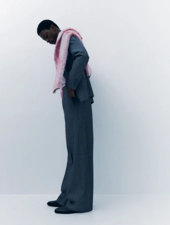 A slender Black male model poses indoors wearing a tailored grey suit, wide-legged trousers, black shoes, and a light pink faux fur stole.