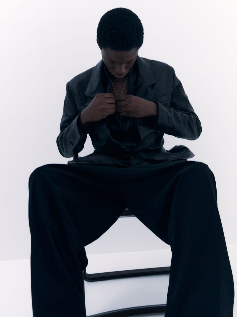 Portrait of a Black male seated on a chair, wearing a grey blazer, and black pants in a bright, minimalist photo studio setting.