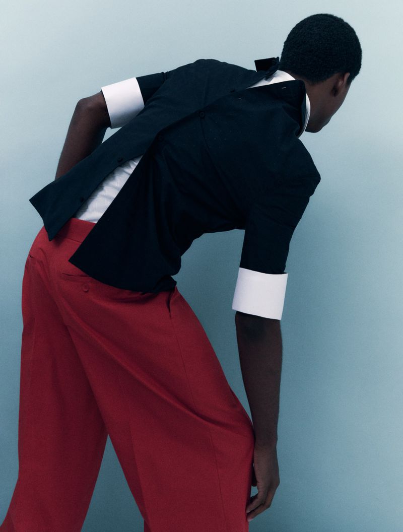 Person poses in black short-sleeve shirt with white collar and cuffs, red pants against a light blue studio backdrop.