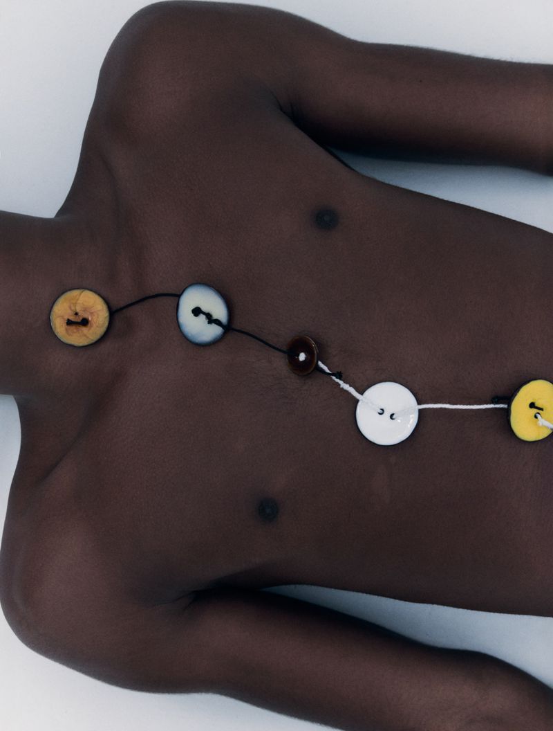 Close-up studio shot of a dark-skinned adult male lying on a white surface, wearing a necklace made of assorted buttons and string.
