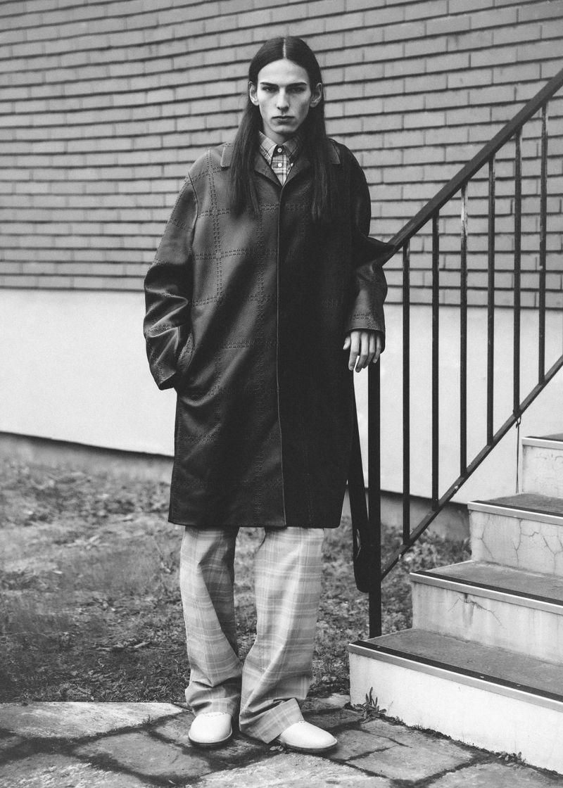Monochrome portrait of a tall, slender adult male with long hair, wearing a dark overcoat, standing on stone steps next to a brick wall.
