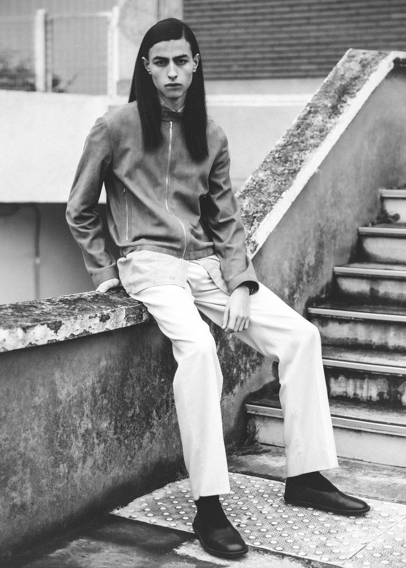 An adult male with long dark hair is seated on a concrete ledge wearing a zip-up jacket, light-colored trousers, and dark shoes in an outdoor setting.