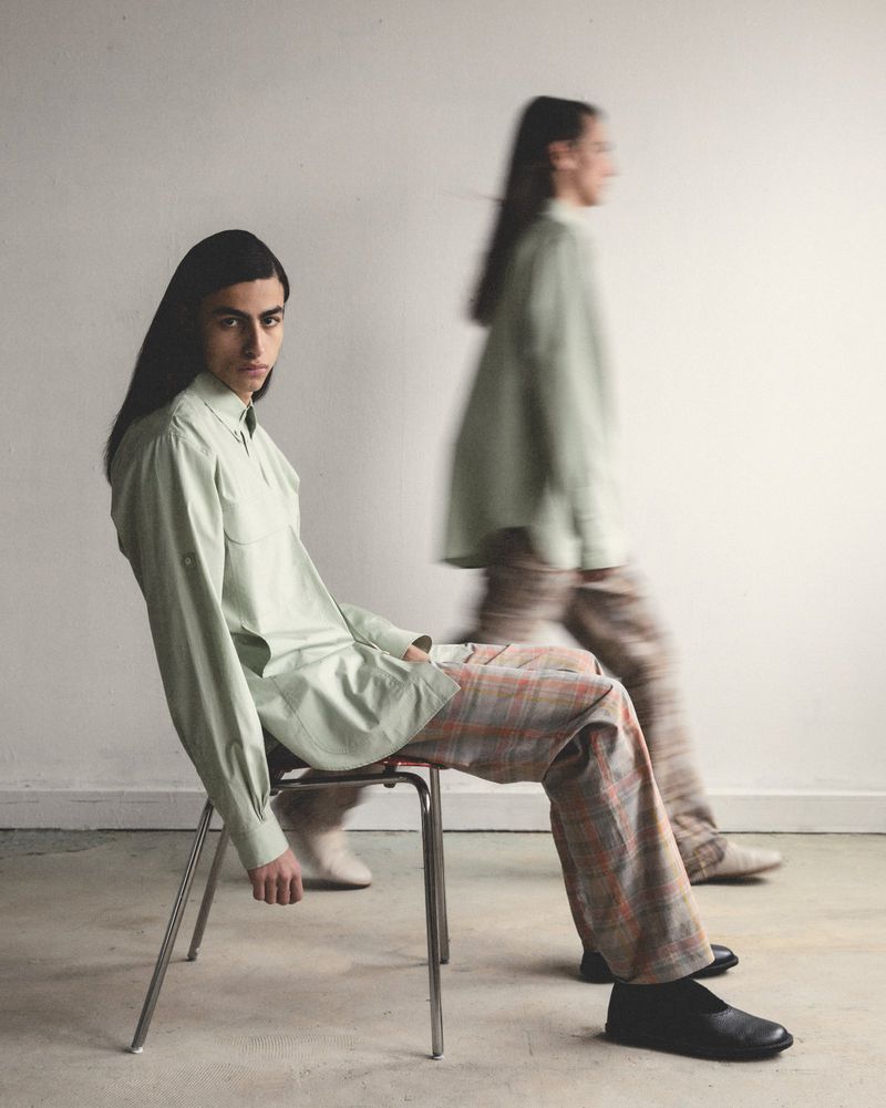 Two people in light green shirts and plaid pants pose indoors; one sits on a metal chair looking at the camera, the other in motion.