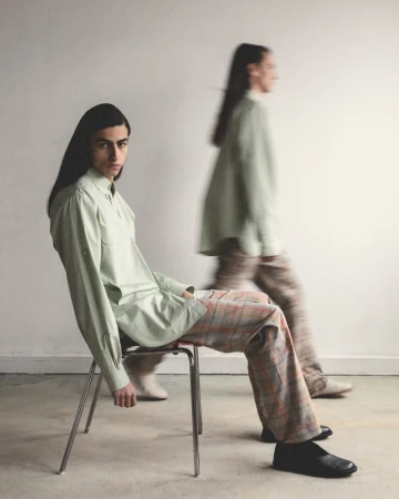 Two people in light green shirts and plaid pants pose indoors; one sits on a metal chair looking at the camera, the other in motion.