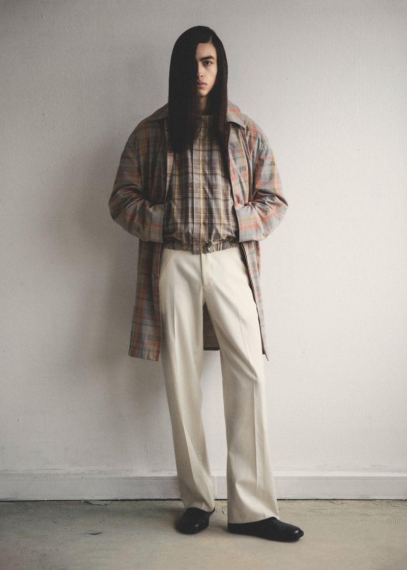 A slender adult male model with long dark hair poses indoors, wearing a long plaid coat, plaid shirt, cream slacks, and black shoes.