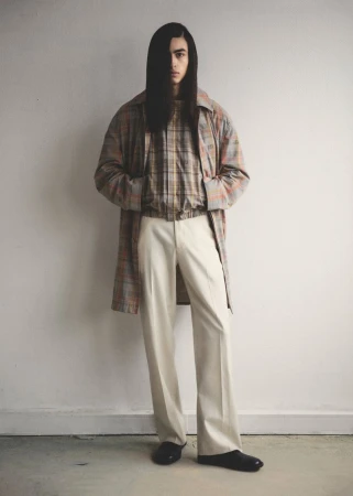 A slender adult male model with long dark hair poses indoors, wearing a long plaid coat, plaid shirt, cream slacks, and black shoes.