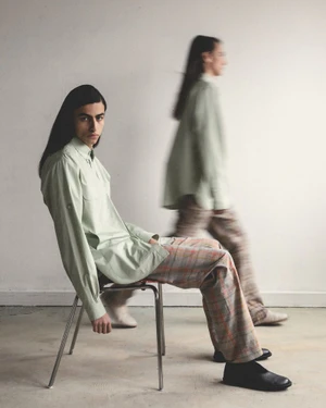 Two male models in HERO Magazine editorial, one seated on a metal chair wearing a sage green shirt and plaid trousers.