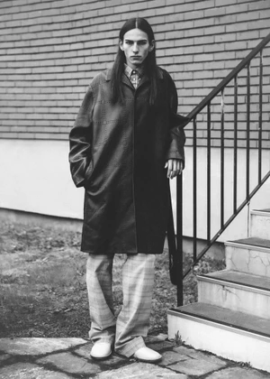 Monochrome fashion portrait of a long-haired male model wearing a dark long coat and patterned trousers near outdoor stairs.