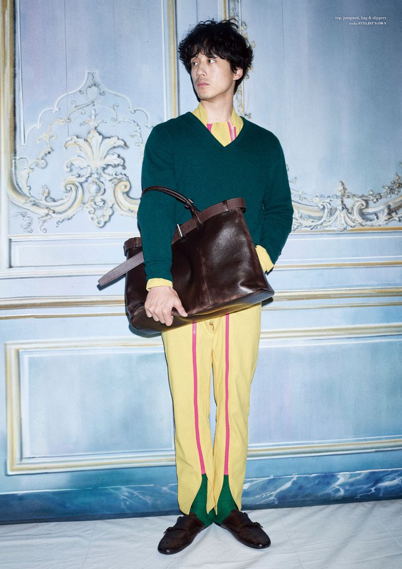Full-length portrait of a slender adult male model in colorful fashion clothing holding a brown leather tote bag, against a patterned wall.