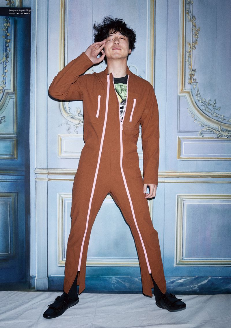 Asian adult male in brown overall suit poses in front of an ornate blue wall with his hand raised to his head.