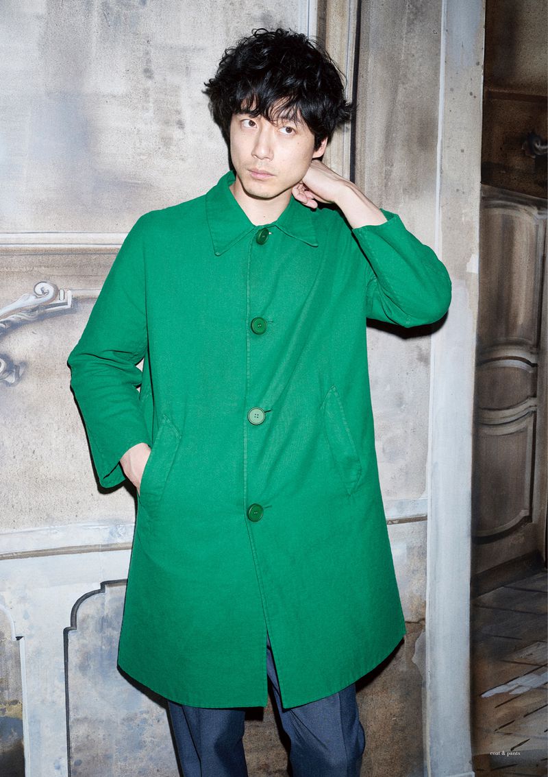 Portrait of a male model in a green coat posing indoors against a decorative wall with muted lighting and a slightly somber mood.