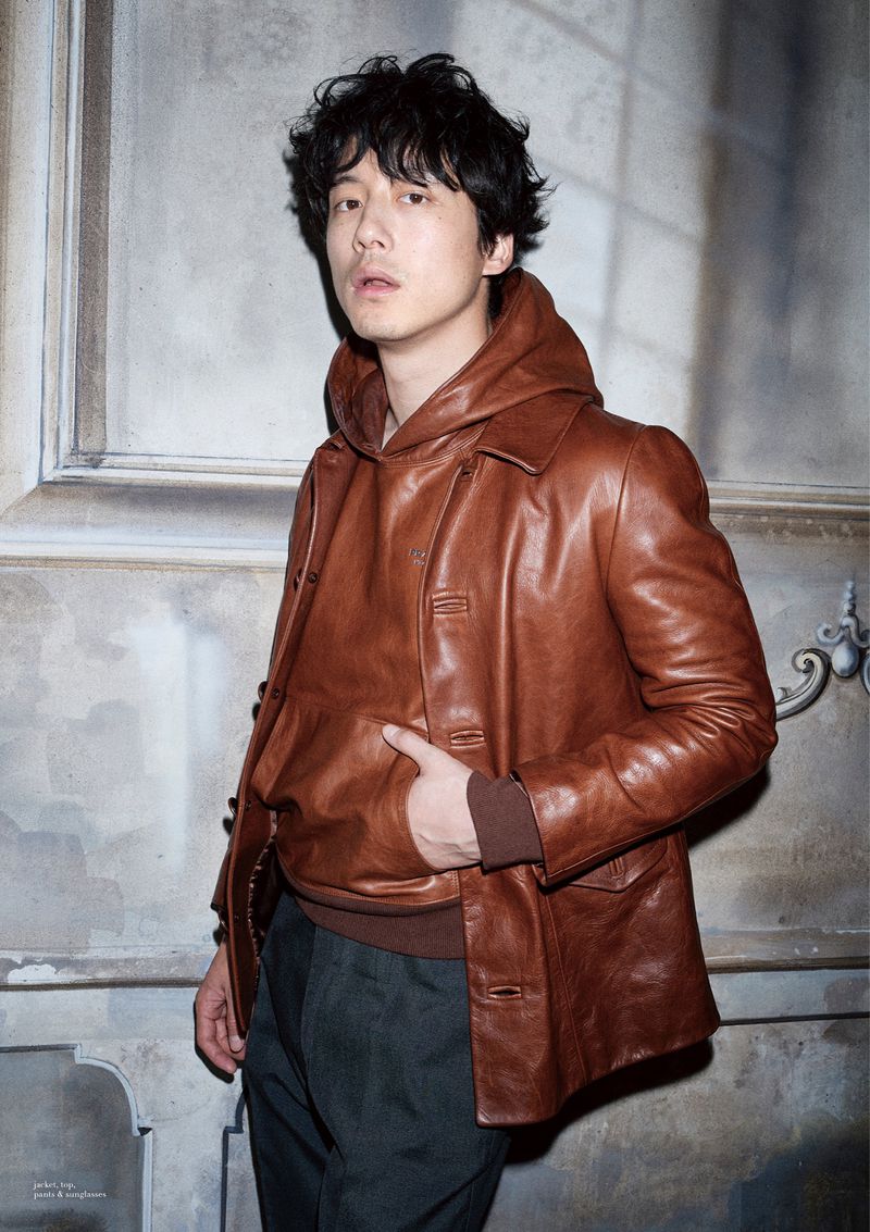 An Asian man stands indoors wearing a brown leather jacket over a hooded sweater, posing in a medium shot against a neutral wall.