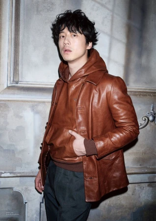 An Asian man stands indoors wearing a brown leather jacket over a hooded sweater, posing in a medium shot against a neutral wall.
