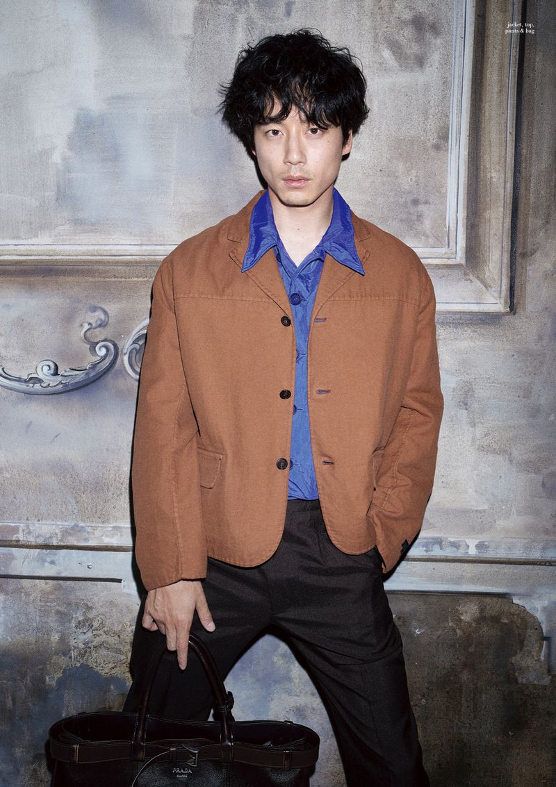 Portrait of an adult male in a tan jacket and blue shirt holding a black tote bag in front of a textured wall.