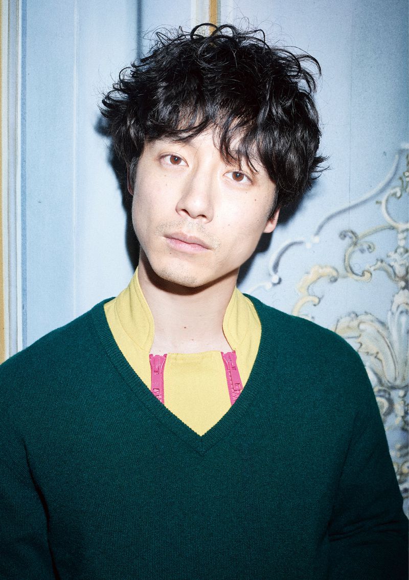 Close-up portrait of a young adult Asian man with dark curly hair wearing a green sweater and a yellow collared shirt indoors.
