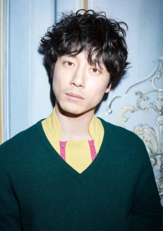 Close-up portrait of a young adult Asian man with dark curly hair wearing a green sweater and a yellow collared shirt indoors.