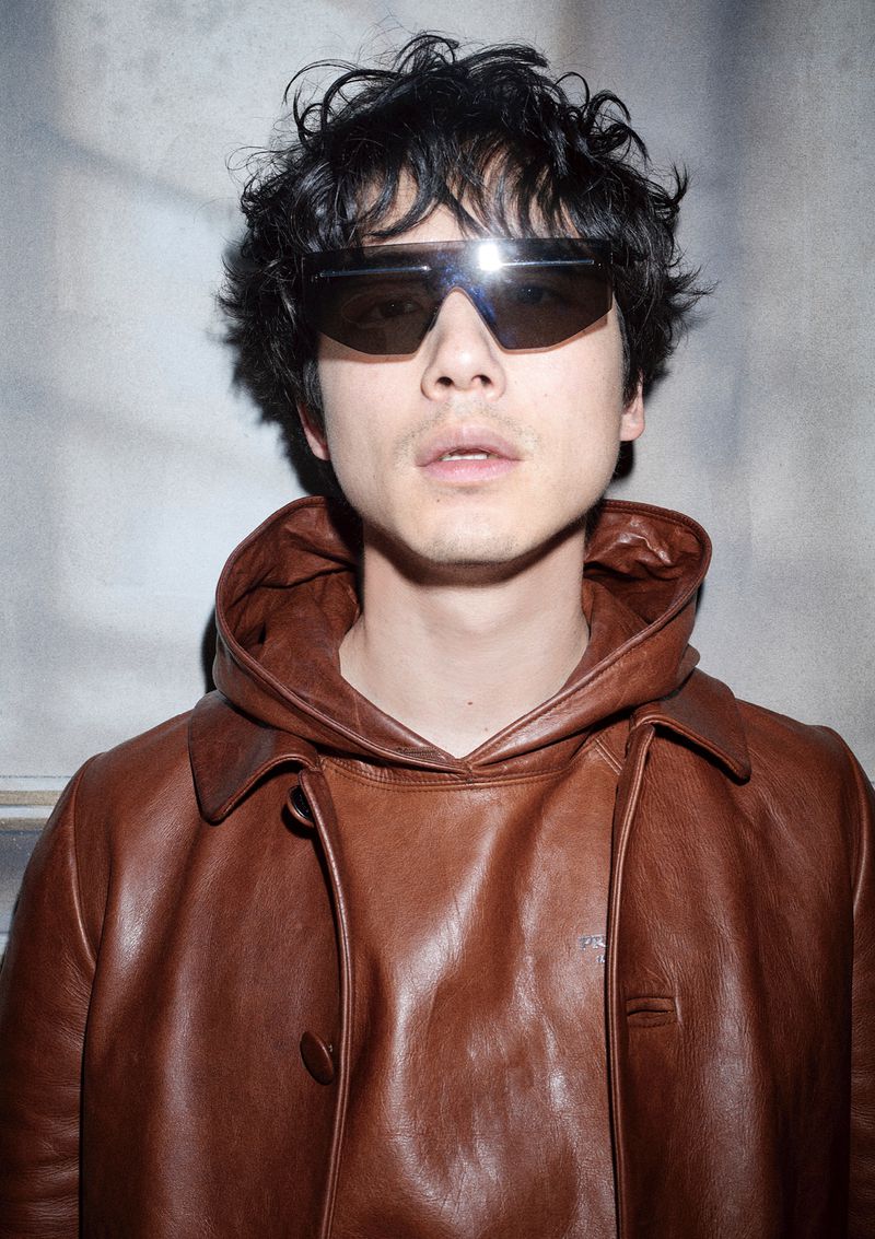 Man wearing dark sunglasses and a brown leather hooded jacket against a textured grey background.