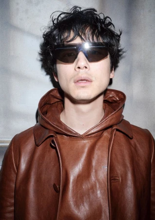 Man wearing dark sunglasses and a brown leather hooded jacket against a textured grey background.