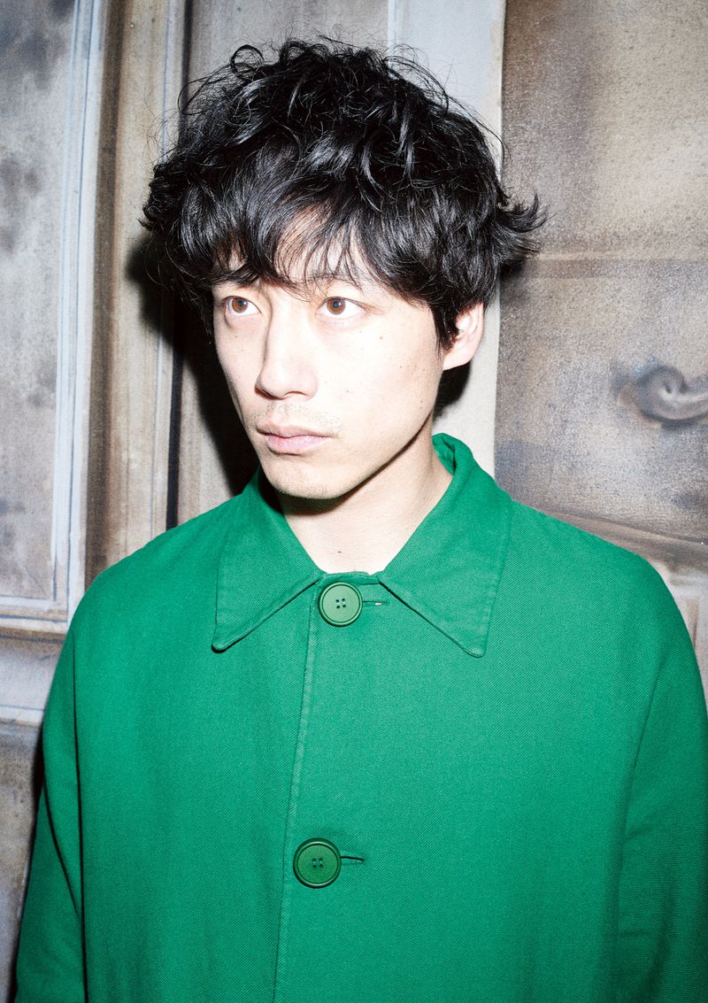 Headshot of an Asian man with black curly hair, wearing a bright green coat, posing in front of an aged stone-like wall in a studio.