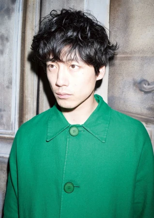Headshot of an Asian man with black curly hair, wearing a bright green coat, posing in front of an aged stone-like wall in a studio.