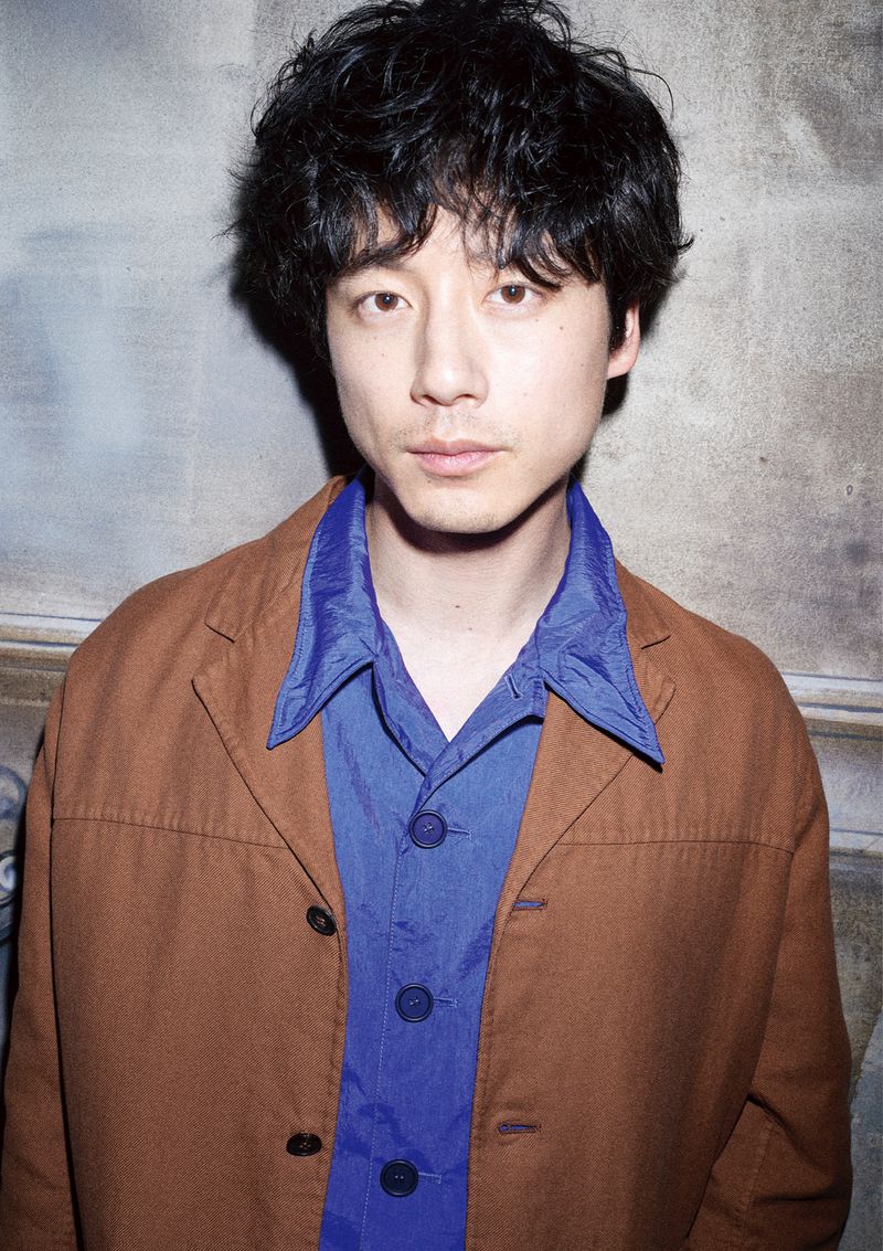 Portrait of a middle-aged Asian man with curly hair wearing a brown jacket over a blue shirt, standing against a textured background.